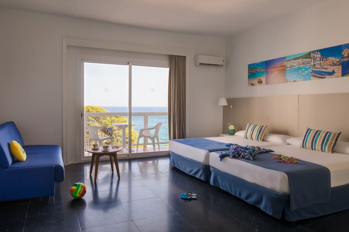 A hotel room with two beds, a balcony with a view of the sea, a small table with a chair, and a blue sofa.