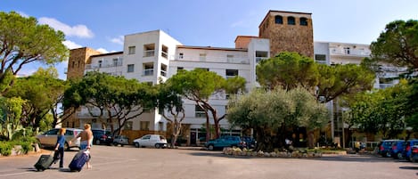 Un estacionamiento con varios coches y personas, rodeado de árboles y edificios con balcones.