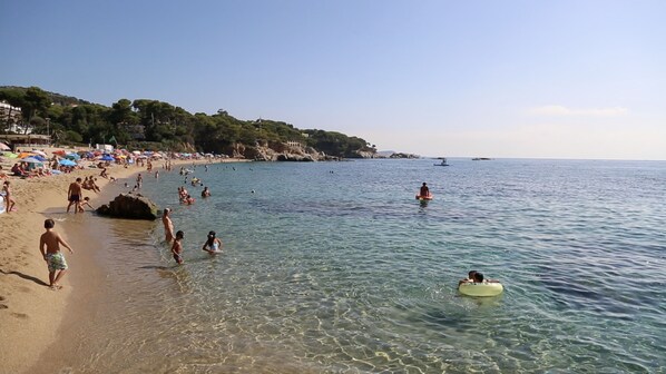 A beach with clear water, people swimming and walking along the shore, and umbrellas set up for sunbathing.