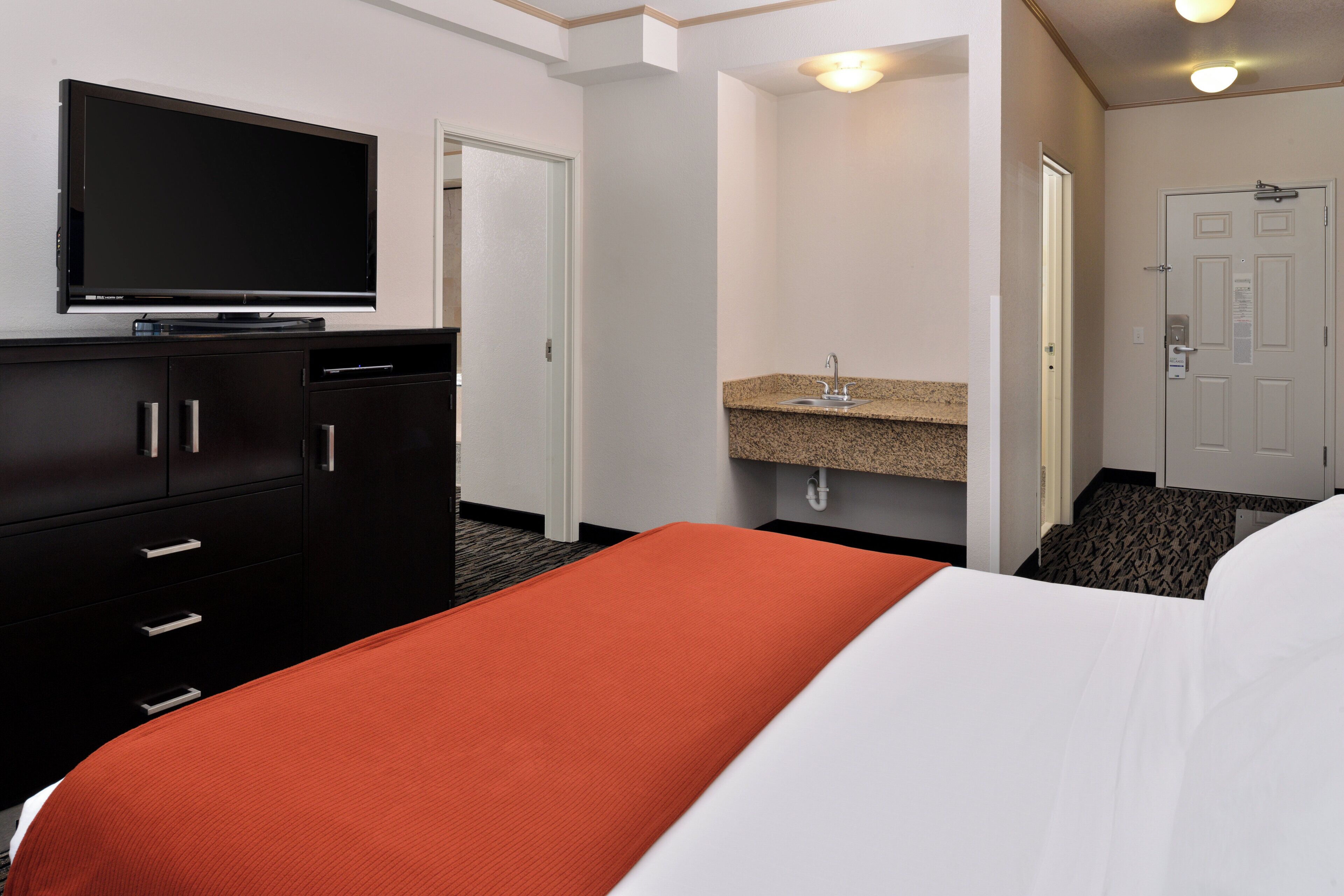 A hotel room with a bed, a television on a dresser, a bathroom with a sink and mirror, and a door.