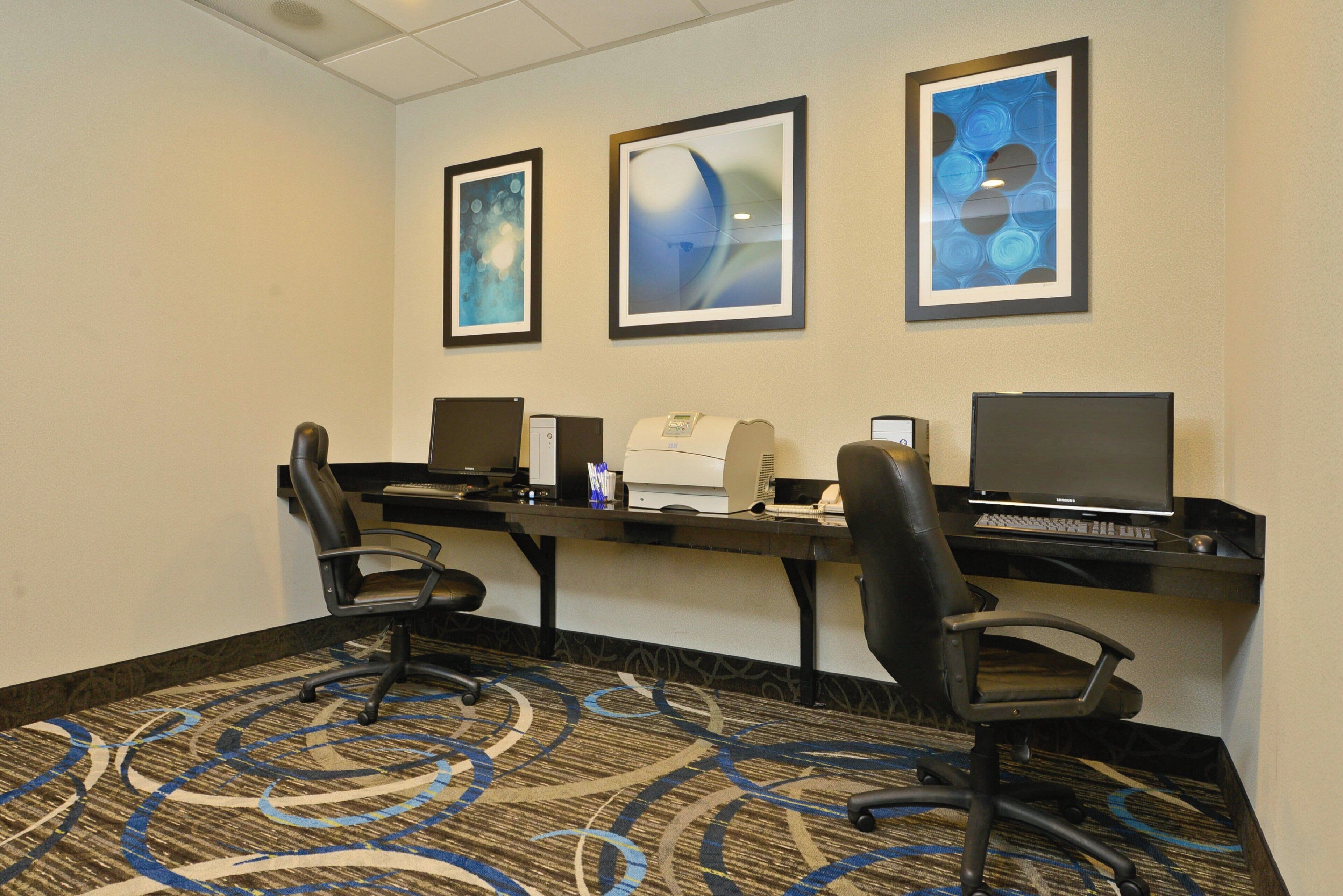 A hotel room with a desk, two chairs, a printer, and three framed artworks on the wall.