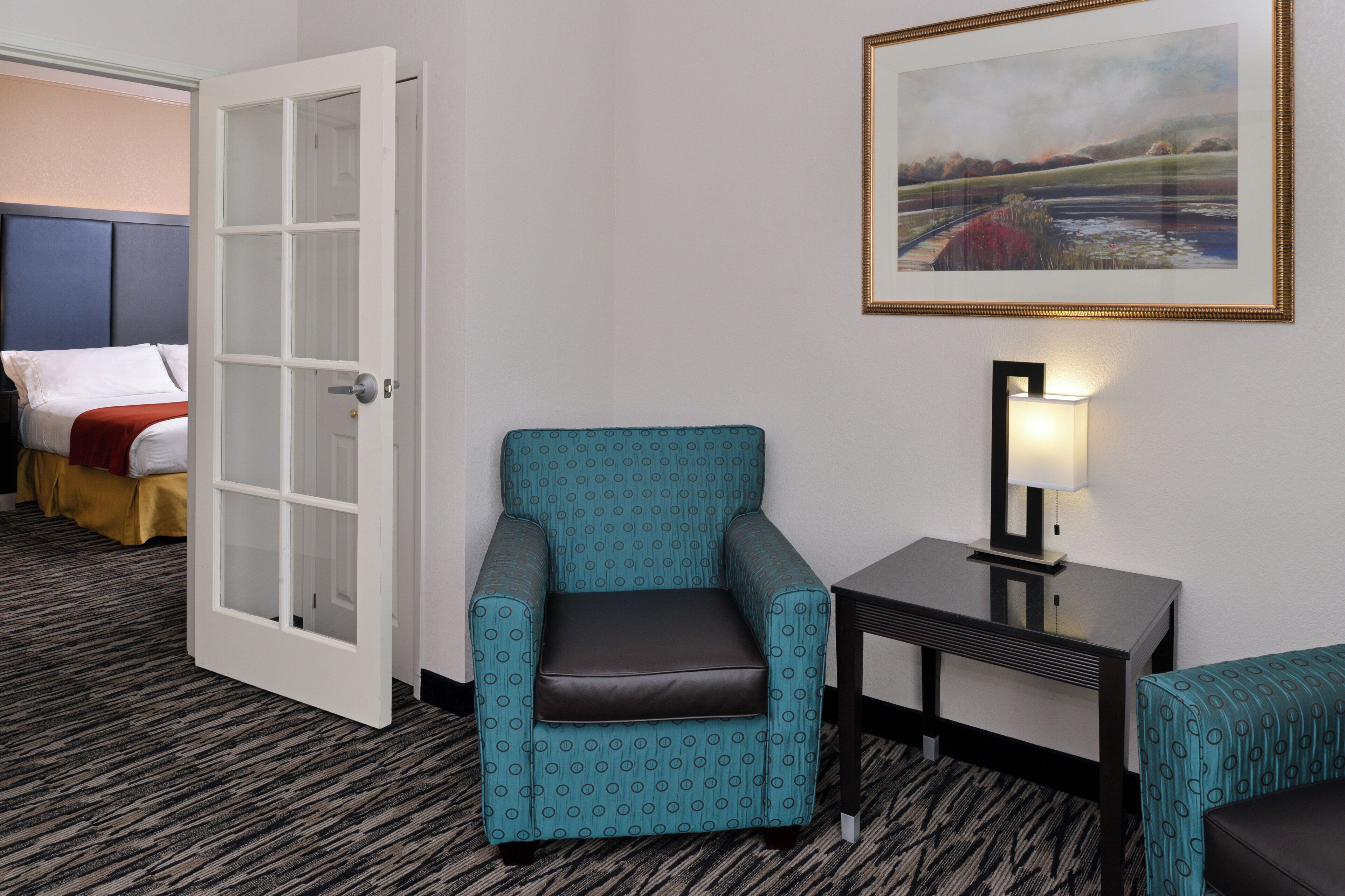 A hotel room with a bed, a blue armchair, a small table with a lamp, and a painting on the wall.