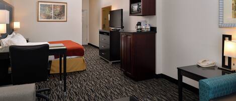 A hotel room with a bed, desk, chair, kitchenette, and a television.