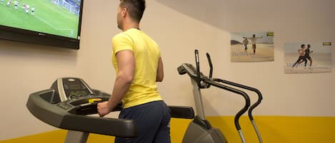 A man walking on a treadmill in a gym with a TV displaying a soccer game and posters on the wall.