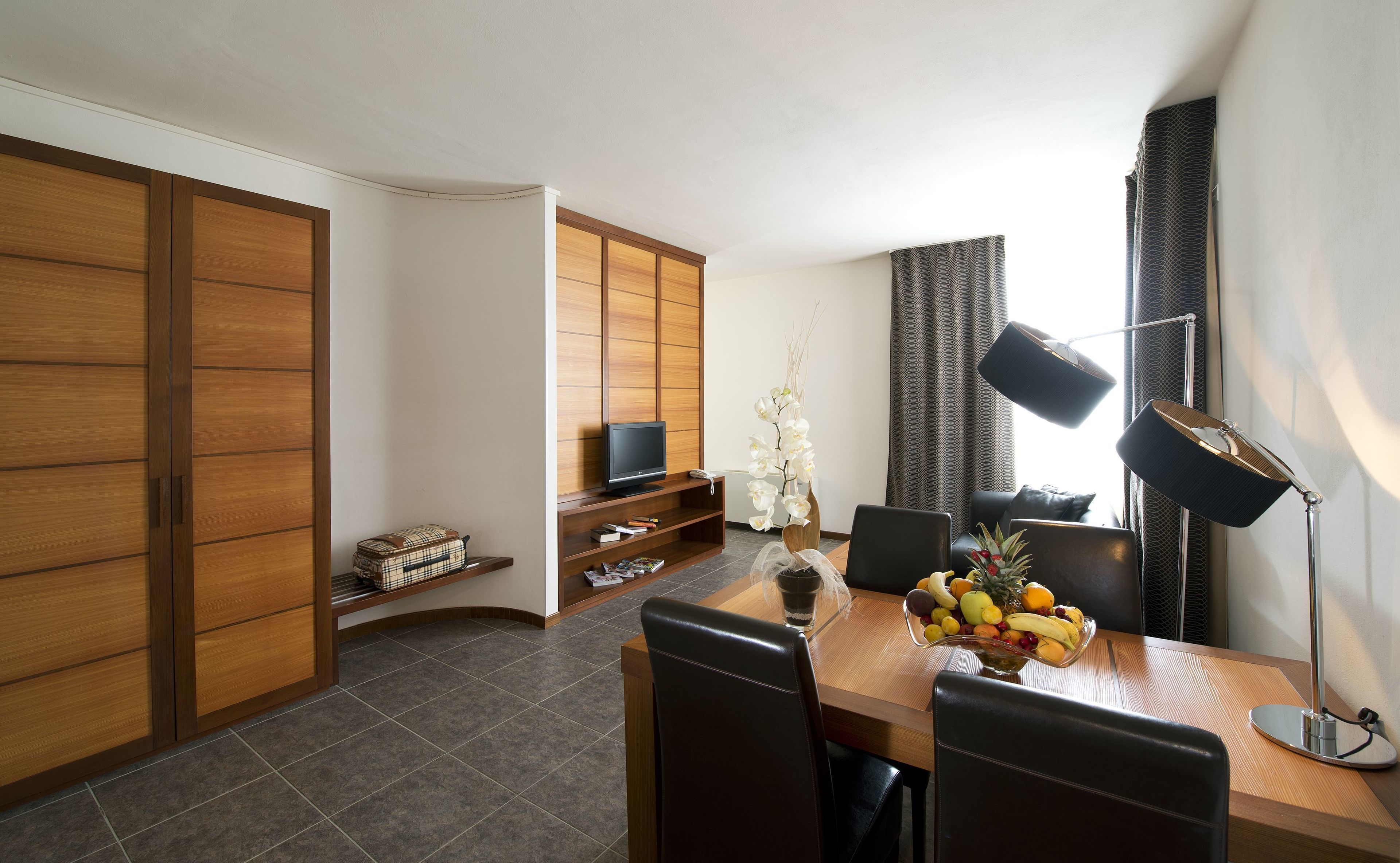 Een moderne woonkamer met een houten vloer, een eettafel met stoelen, een televisie, een boekenkast en een groot raam met gordijnen.