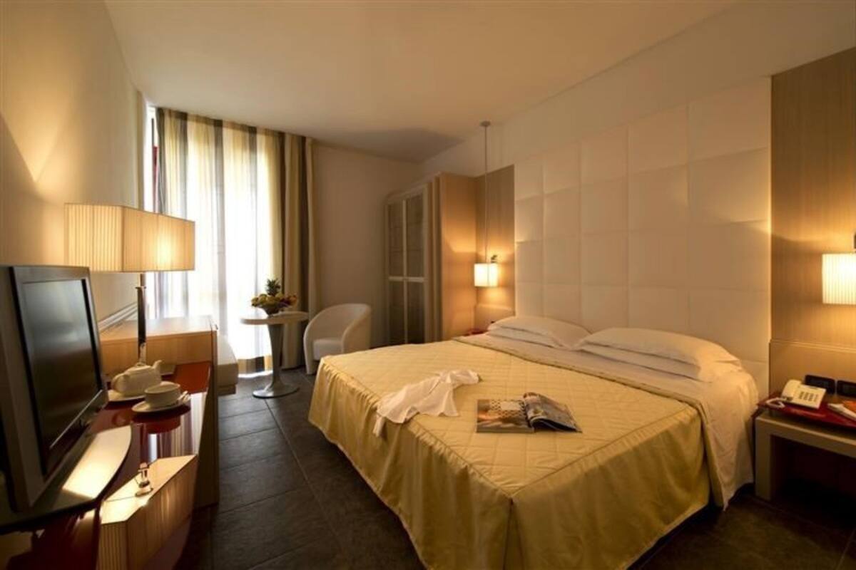 Habitación de hotel con cama, mesita de noche, lámpara, sillón y escritorio con televisor.