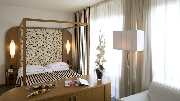 Ein Hotelzimmer mit Himmelbett, Nachttisch mit Zeitschrift und Früchten, Lampe und Blumen in einer Vase.