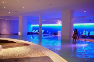 Una piscina interior con iluminación azul, sillas de descanso y una persona de pie junto a la piscina.