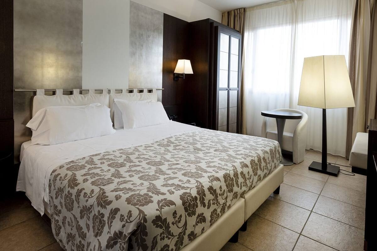Habitación de hotel con una cama grande, una mesita de noche, una lámpara y vistas al exterior a través de la ventana.