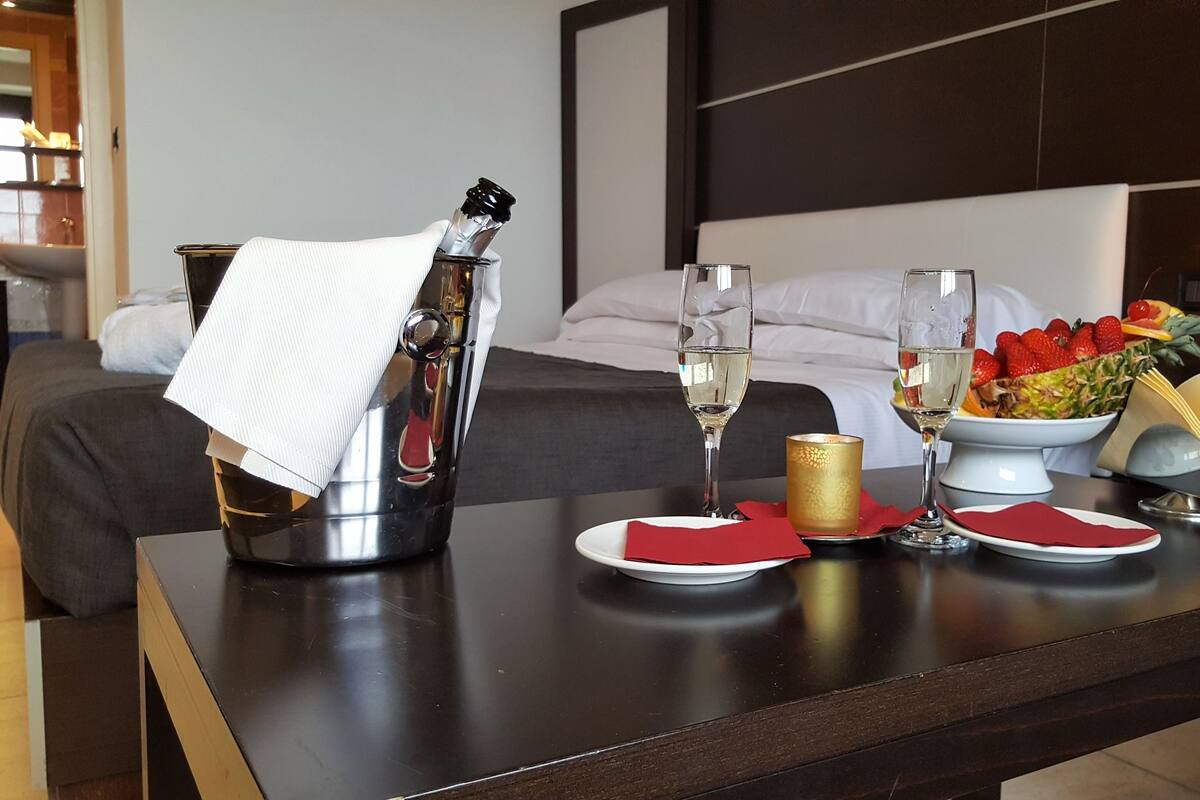 Habitación de hotel con cama, mesita de noche con una botella de champán, dos copas de champán, frutero y una vela.