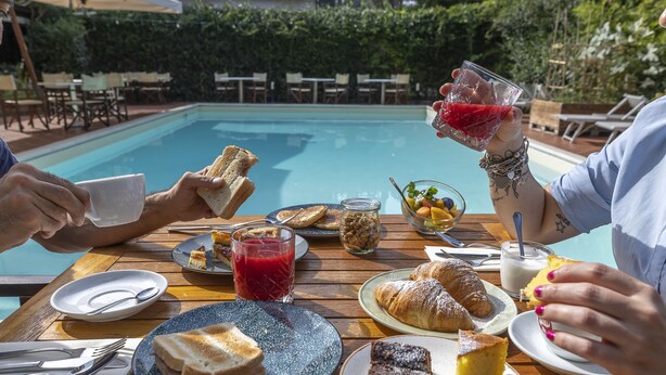 Una mesa de comedor con platos de comida, un vaso de bebida roja y una piscina al fondo.
