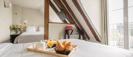Habitación de hotel con cama, bandeja de desayuno, periódico y vistas a la ciudad a través de amplias ventanas.