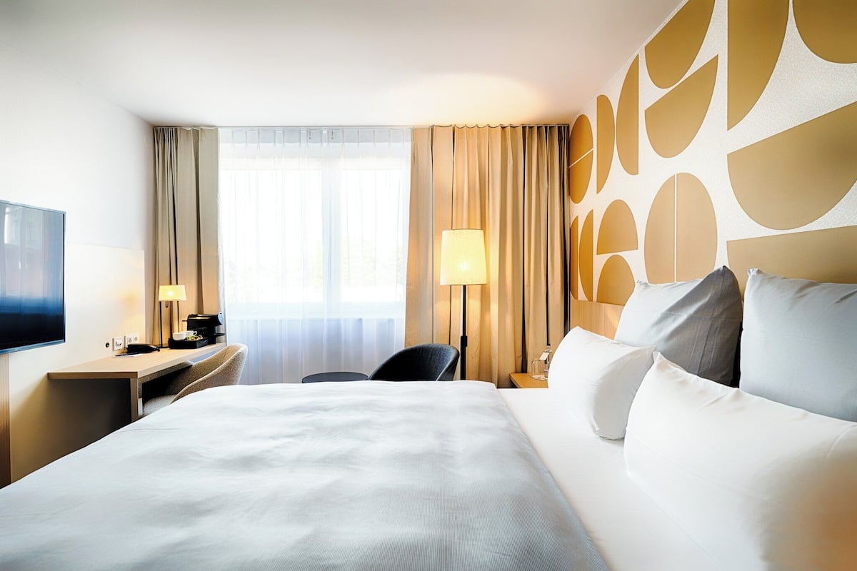 Ein Hotelzimmer mit einem großen Bett, einem Schreibtisch mit Lampe, einem Sessel und einem Fernseher.