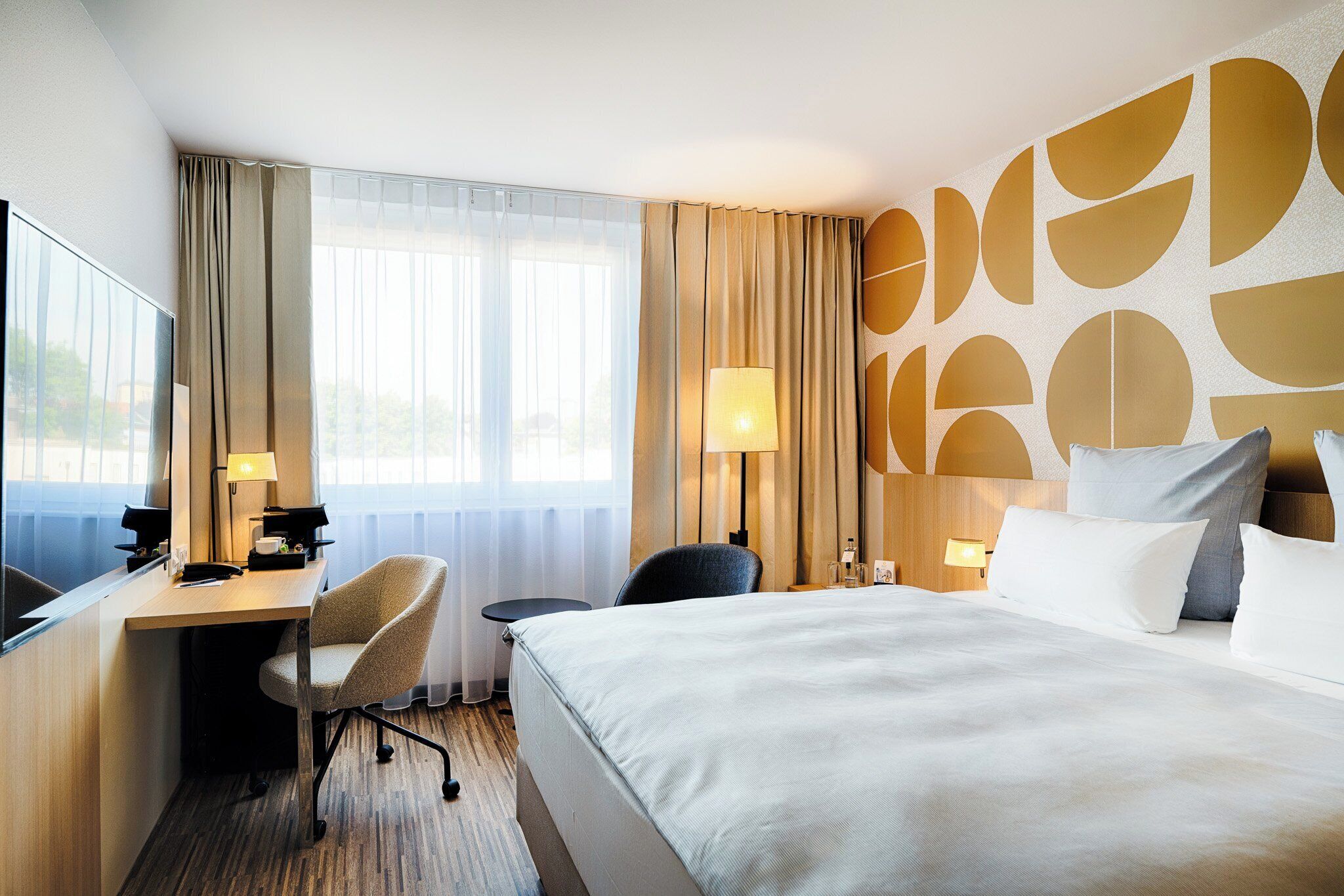 Ett hotellrum med en säng, ett skrivbord, en stol och ett stort fönster med gardiner.