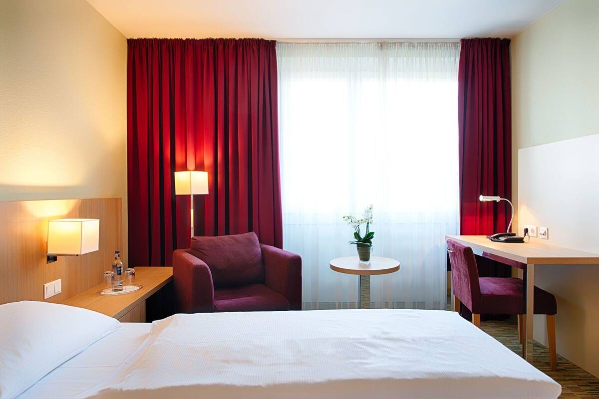 Ein Hotelzimmer mit einem Bett, zwei Stühlen, einem Schreibtisch, einer Lampe, einem kleinen Tisch mit Pflanze und Vorhängen.