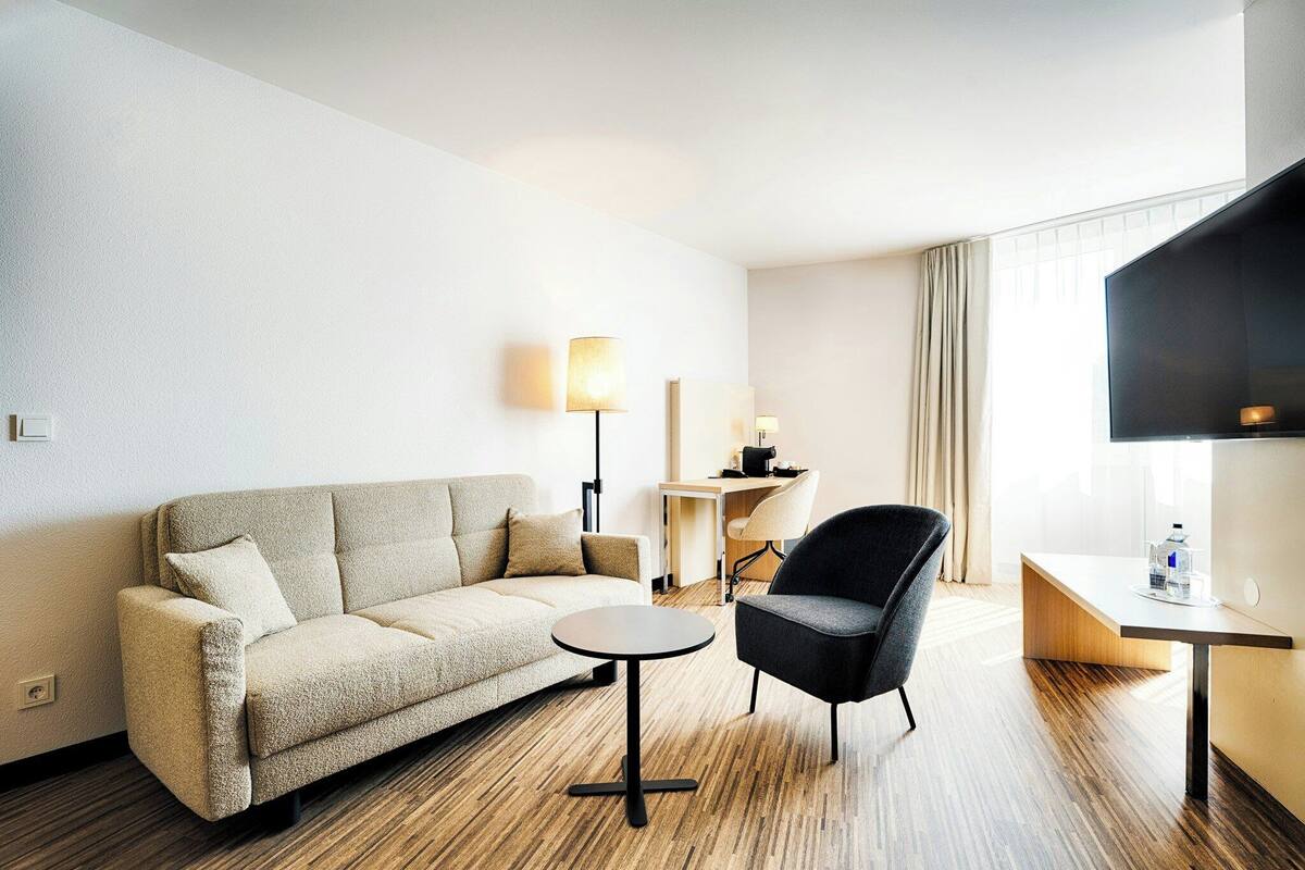 Ein Hotelzimmer mit Sofa, Sessel, kleinem Tisch, Lampe, Schreibtisch und Fernseher.