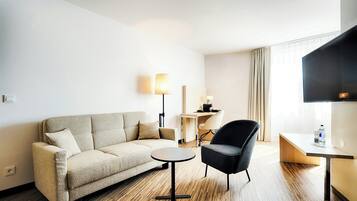 Ein Hotelzimmer mit Sofa, Sessel, kleinem Tisch, Lampe, Schreibtisch und Fernseher.