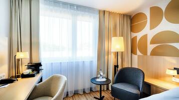 A hotel room with a desk, two chairs, a small table with a lamp, and a bed.