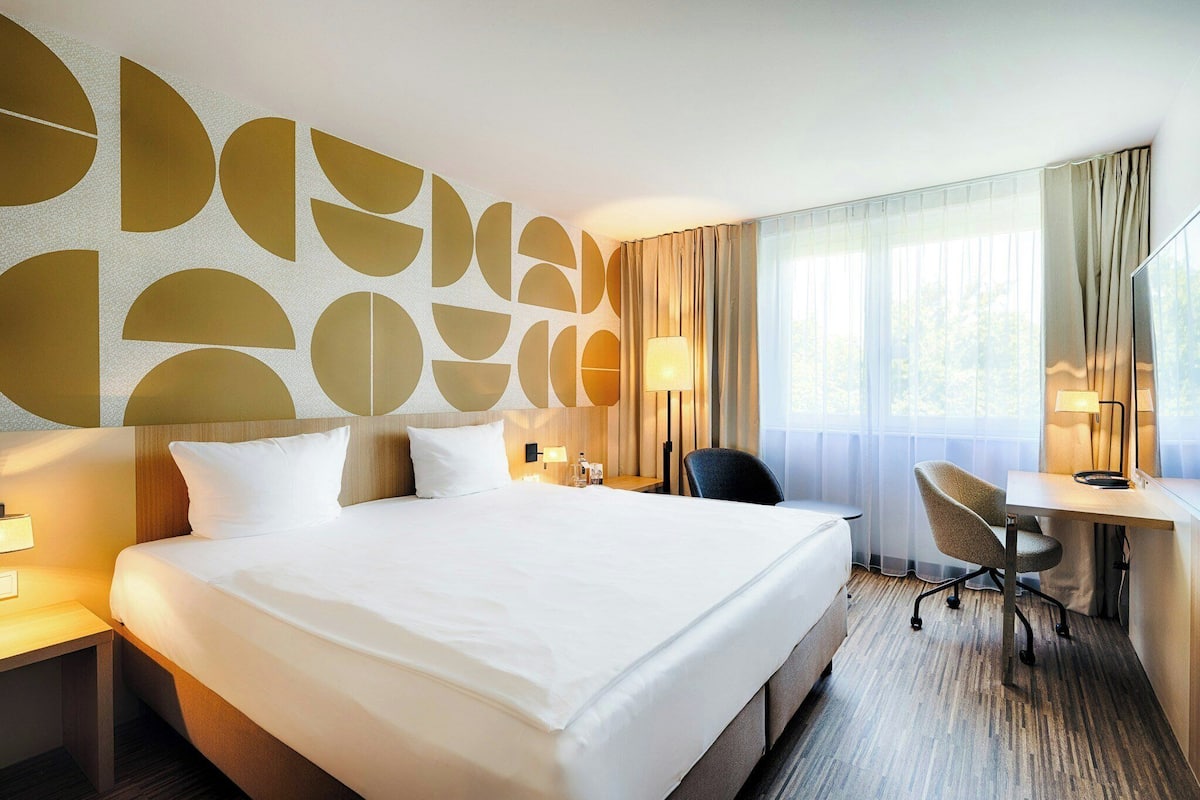Ein Hotelzimmer mit einem großen Bett, einem Schreibtisch und einem Stuhl. Es gibt ein Fenster mit Vorhängen, eine Wand mit geometrischem Muster und eine Lampe auf dem Schreibtisch.