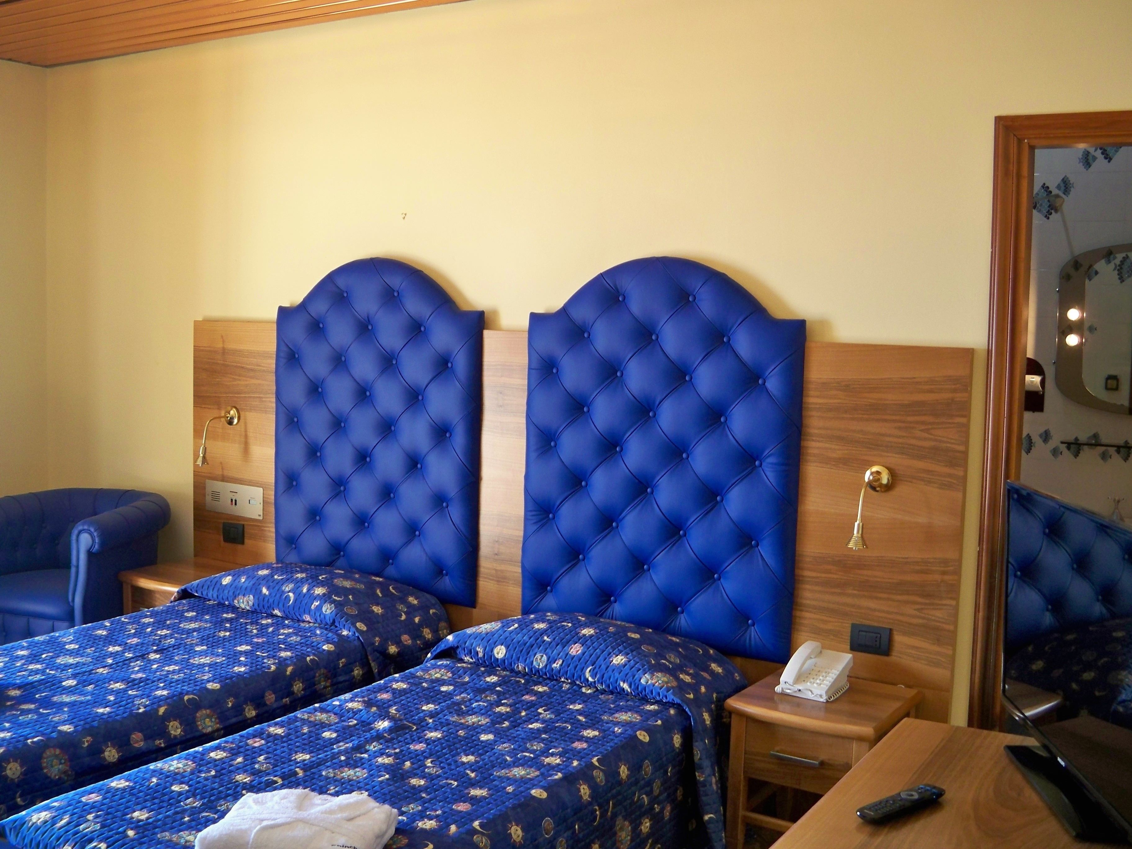A hotel room with two beds, each with a blue headboard and matching bedding. There is a wooden nightstand with a telephone and a remote control on it.