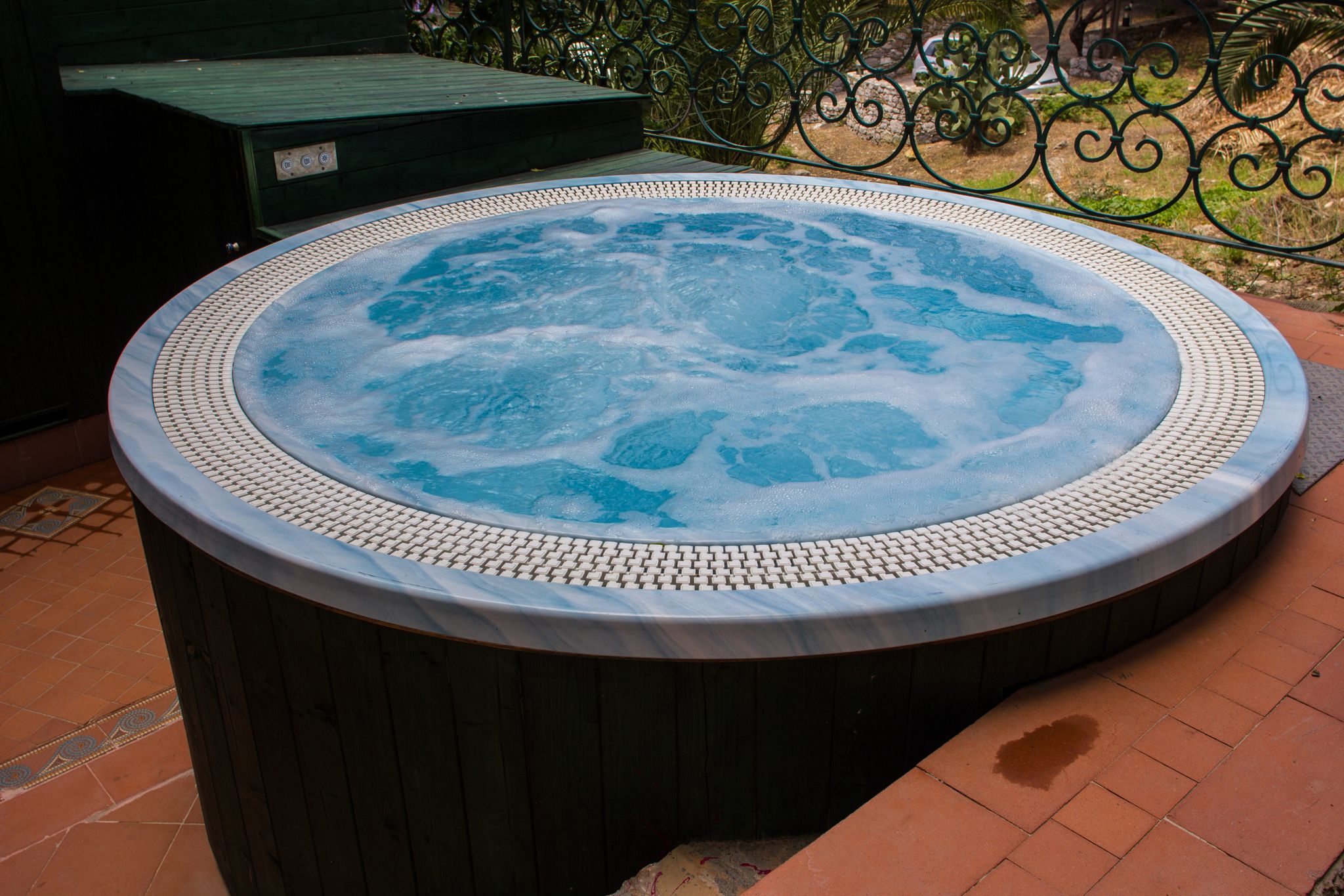Un bain à remous bordé de carreaux, entouré d’une clôture en métal et d’une terrasse en bois.