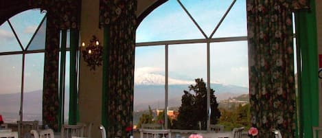 Un coin repas avec des tables préparées pour le service du thé, de grandes fenêtres offrant une vue sur les montagnes et des rideaux à motifs floraux.