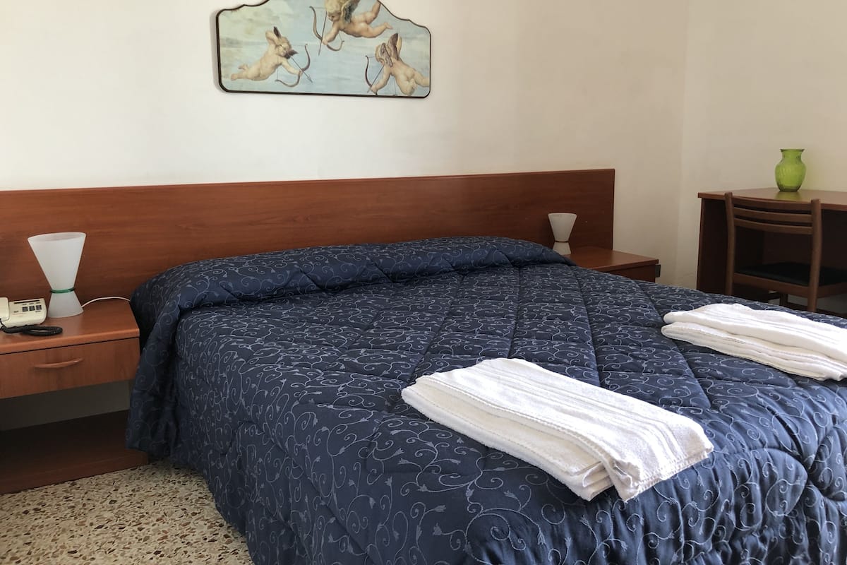 A neatly made bed with a blue patterned duvet, two bedside lamps, a green vase, and a wall painting of cherubs.