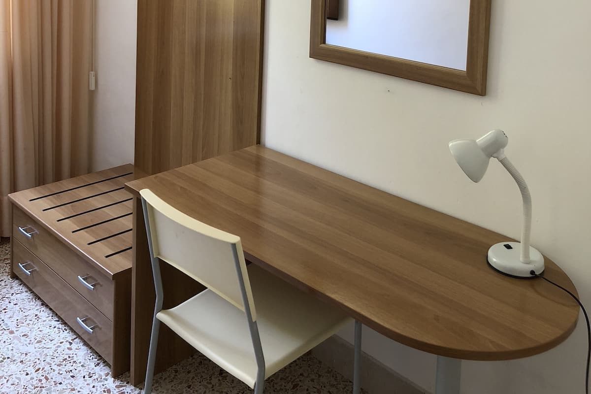 A compact room with a wooden desk, a chair, a desk lamp, a wardrobe, and a framed picture on the wall.