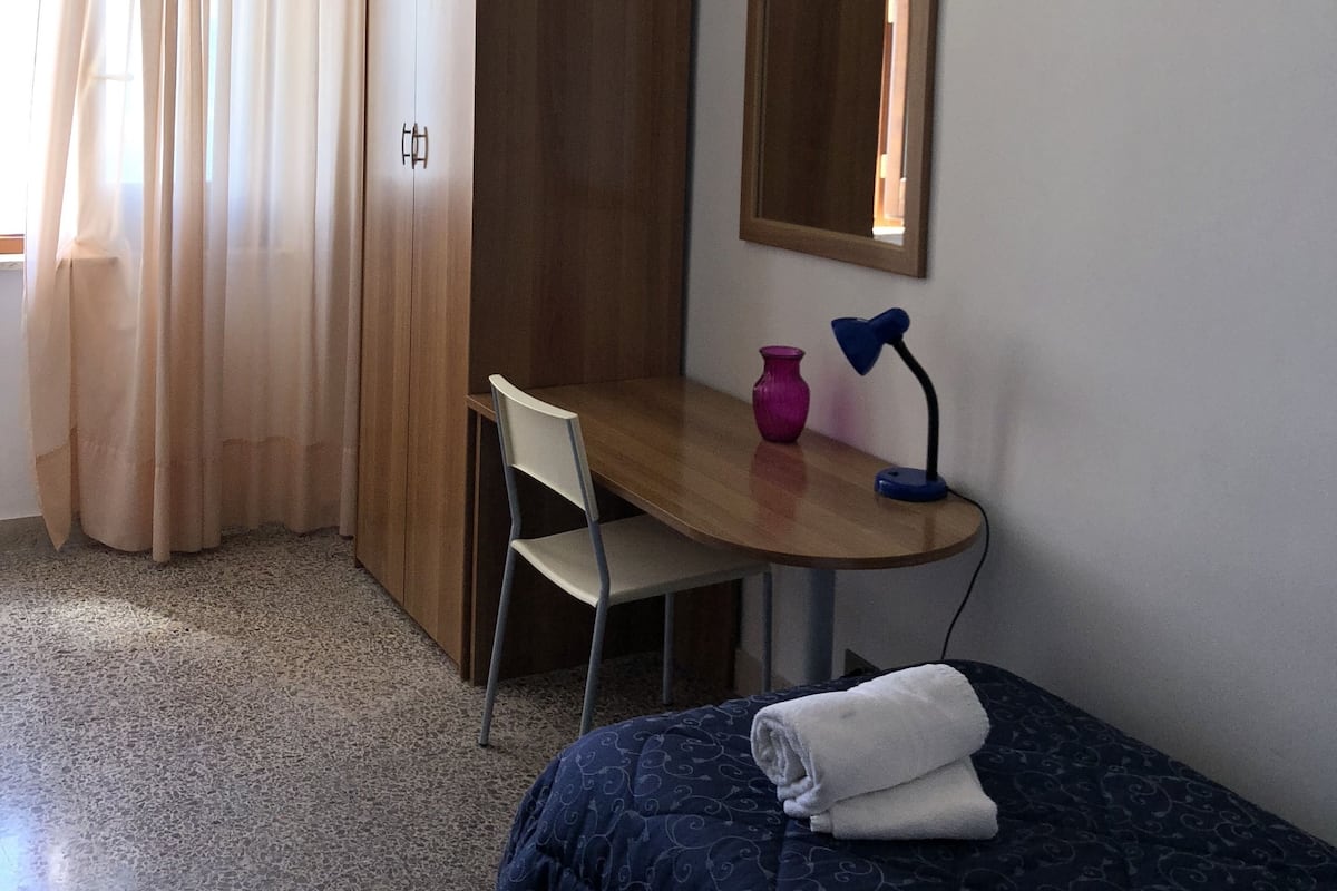 A bedroom with a bed, a desk, a chair, a wardrobe, a mirror, and a window with curtains.