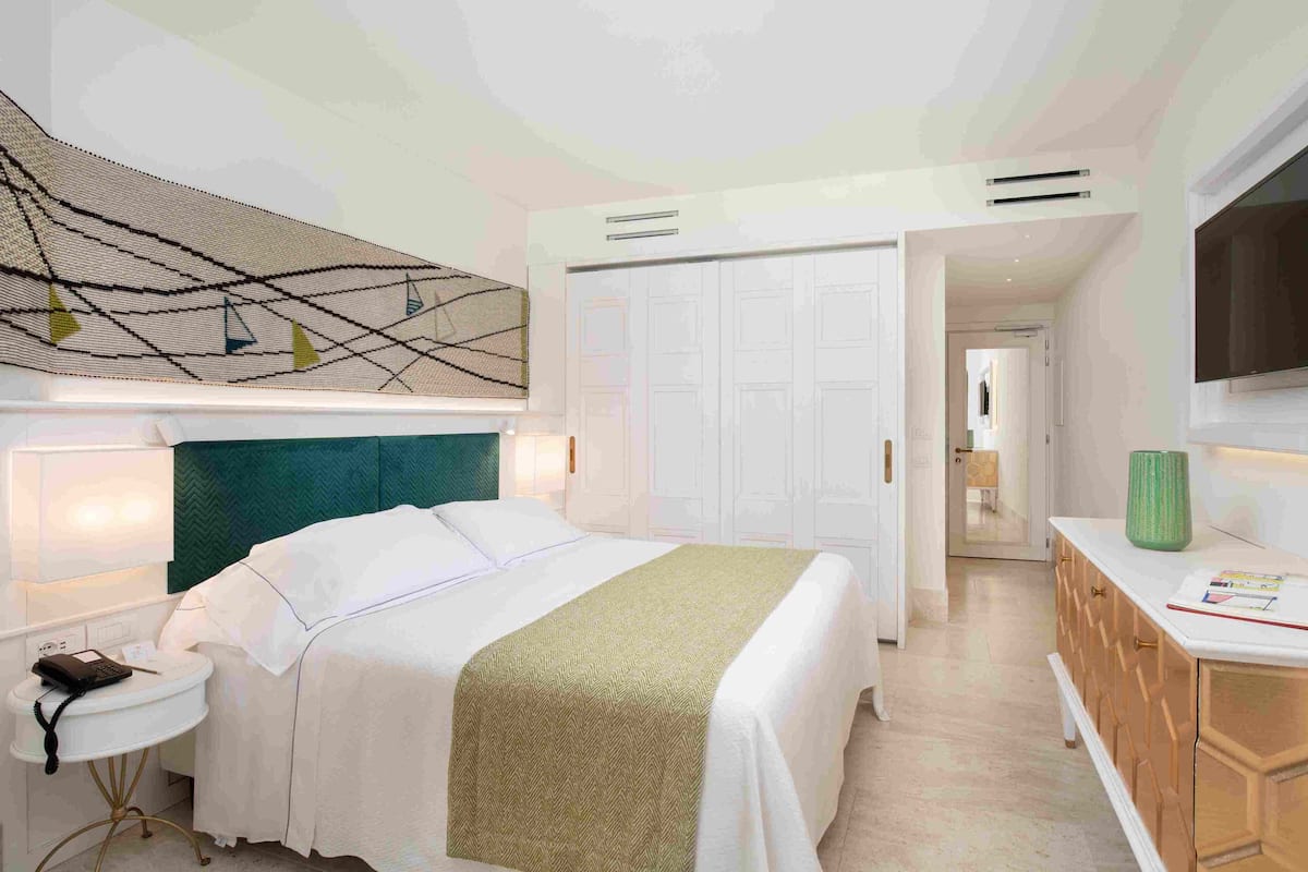 A hotel room with a bed, bedside table, lamp, wardrobe, and a television on the wall.