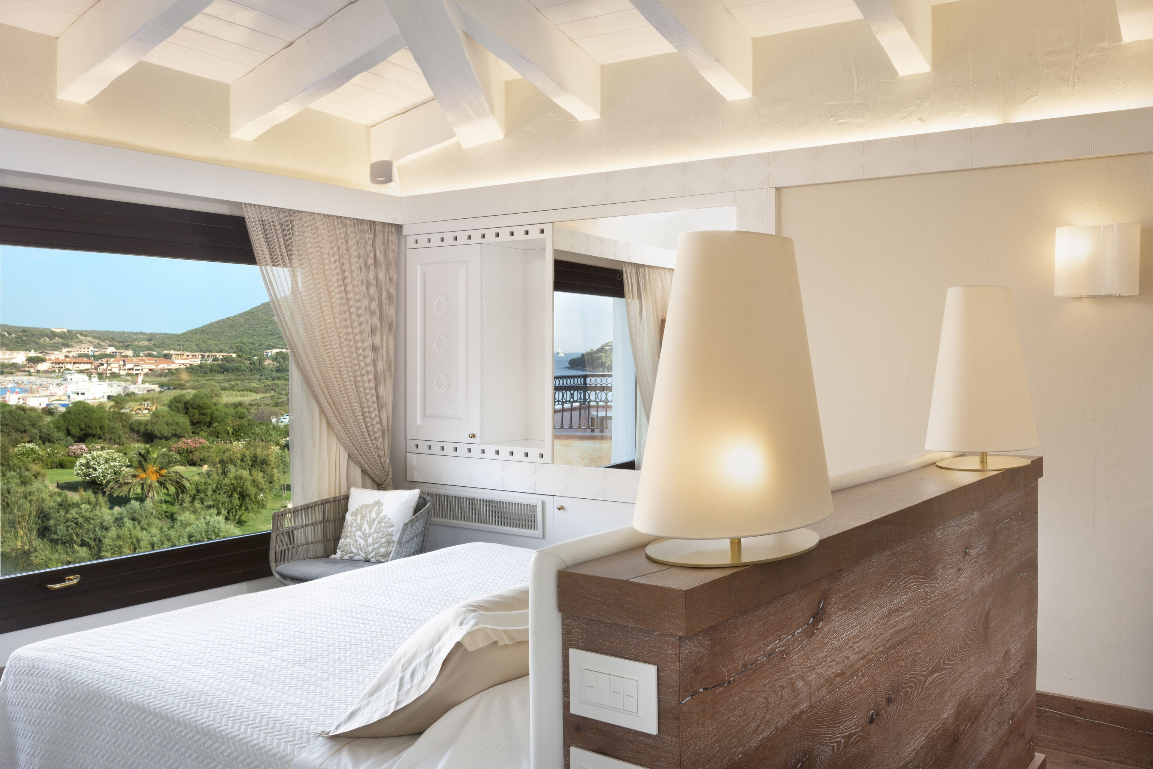 Une chambre à coucher avec un grand lit, une tête de lit en bois, une table de chevet avec des lampes, un miroir et une vue sur un paysage verdoyant par la fenêtre.