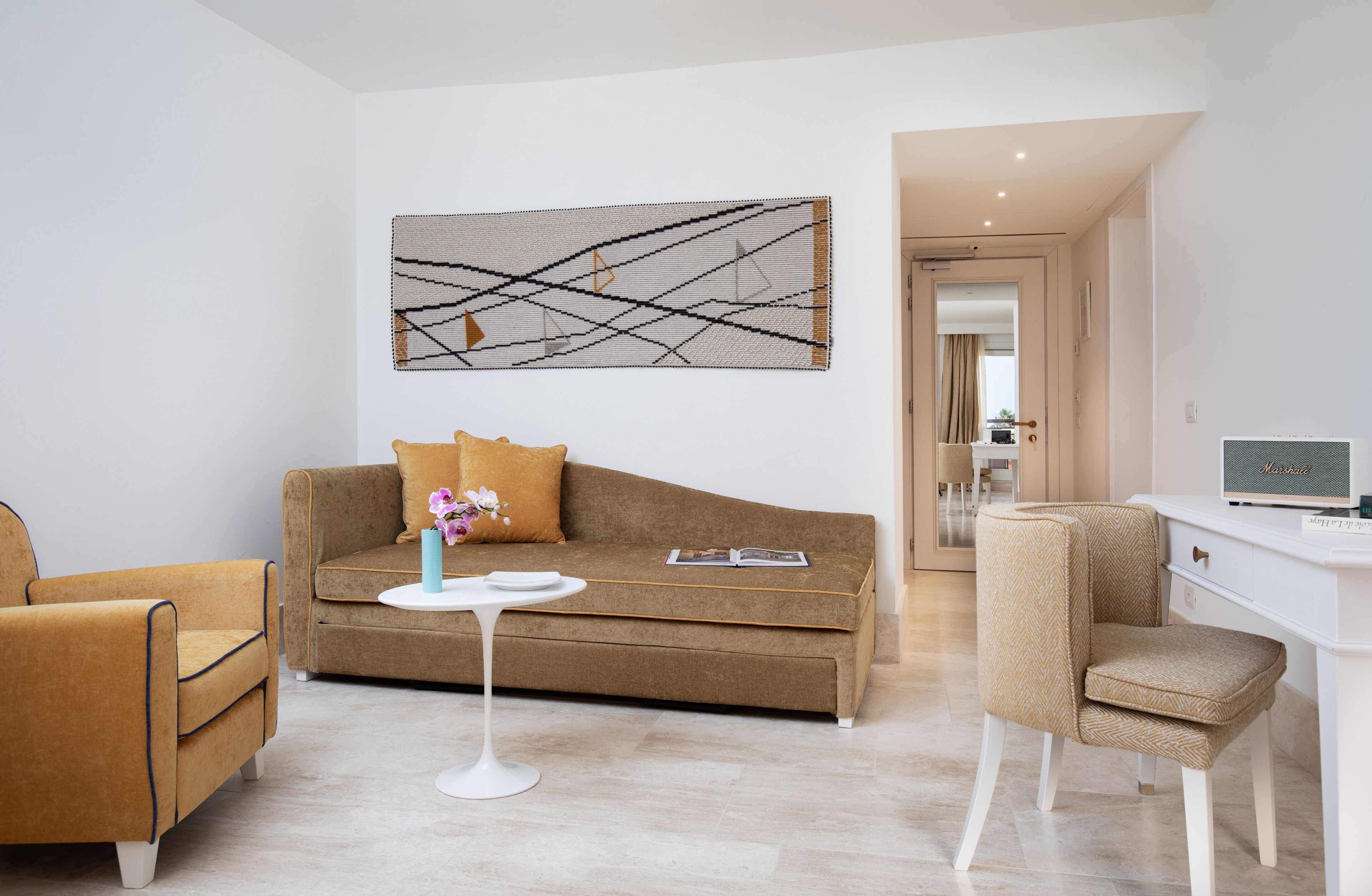 A modern living room with a brown sofa, a white coffee table, and a beige armchair. A large abstract painting hangs on the wall, and a laptop is on a white desk.