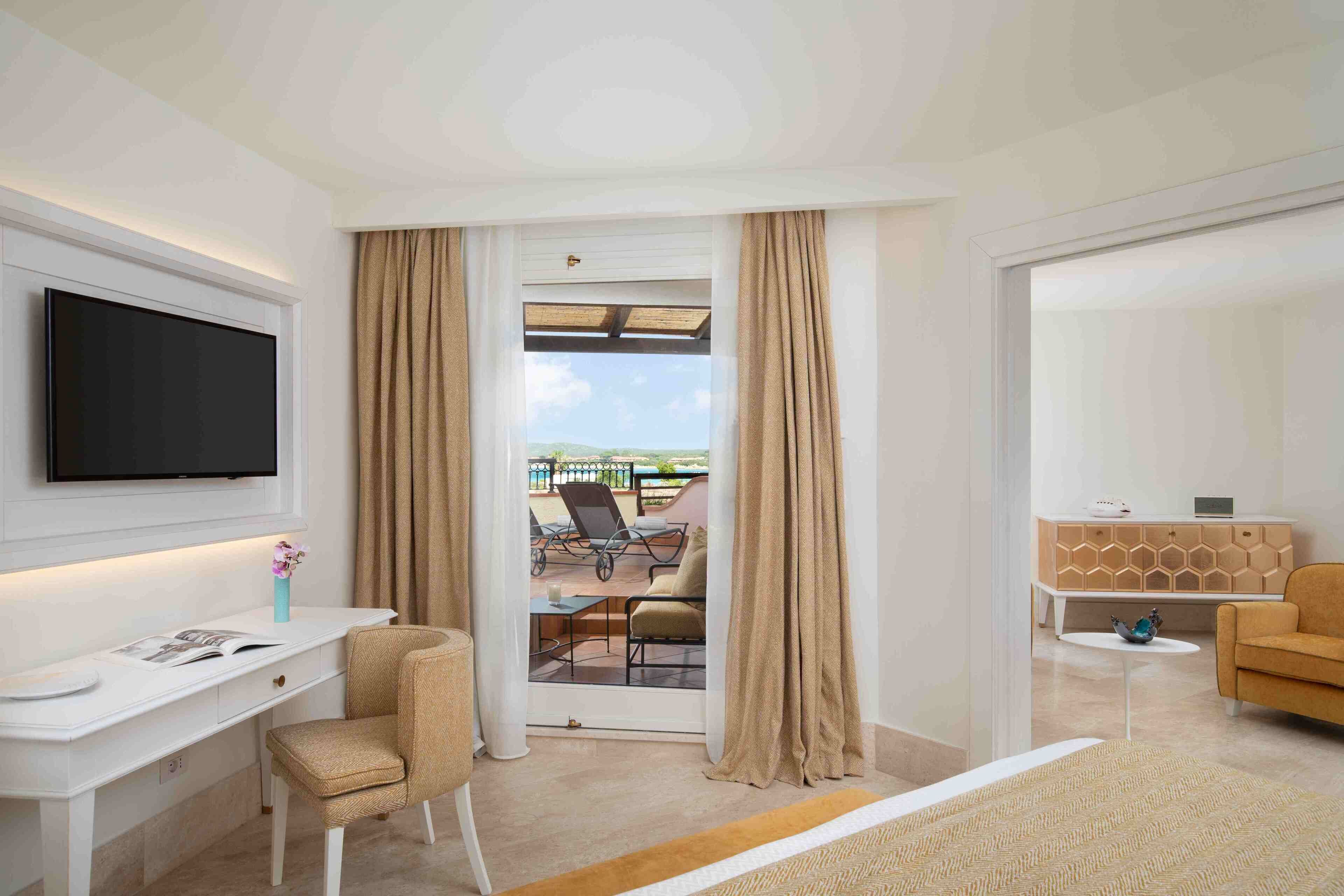 Ett hotellrum med en platt-TV som är monterad på väggen, ett skrivbord med en stol, en säng med ett beige överkast, ett litet bord med en dekorativ skål och utsikt över den utomhusavdelning som utgörs av balkongen.