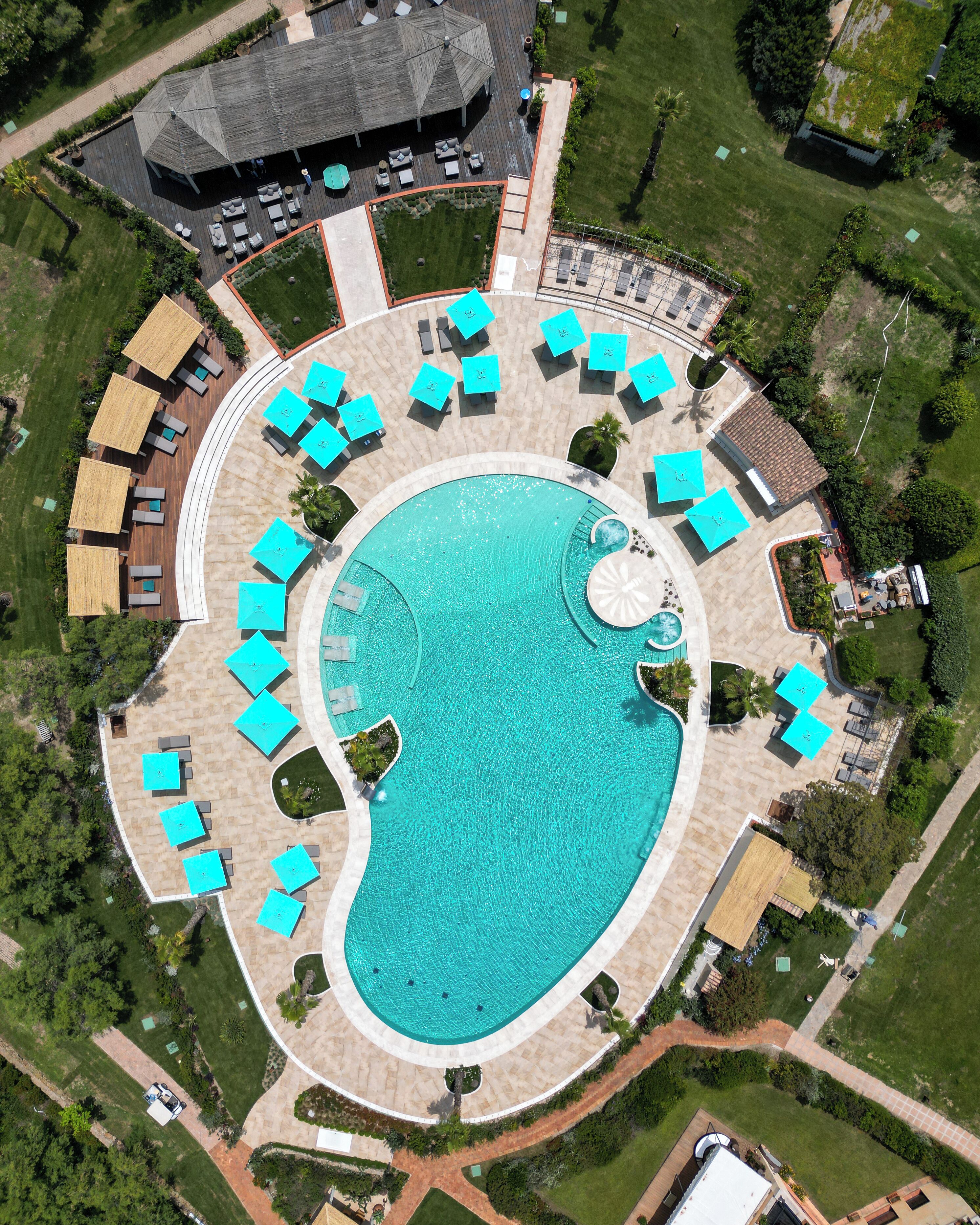 Ett flygbildsfoto av ett semesterområde med en stor pool i mitten, solstolar och en täckt utomhusplats med sittmöbler.