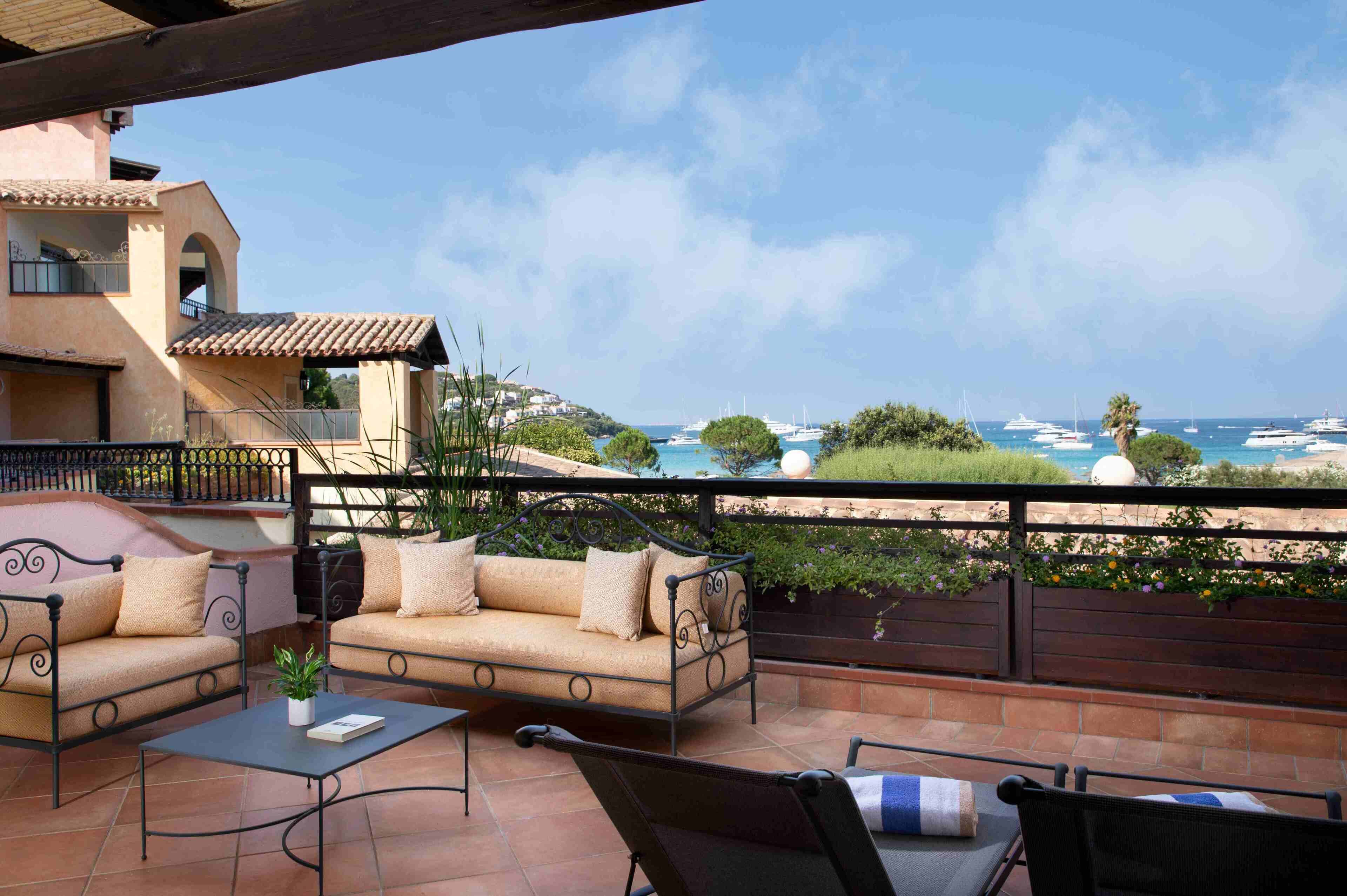 A terrace with a sofa, chairs, and a table, overlooking a coastal area with boats and greenery.