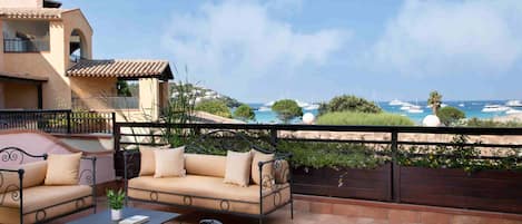 A terrace with a sofa, chairs, and a table, overlooking a coastal area with boats and greenery.