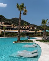 Piscina de resort con palmeras, un elemento acuático y edificios de estilo mediterráneo al fondo.