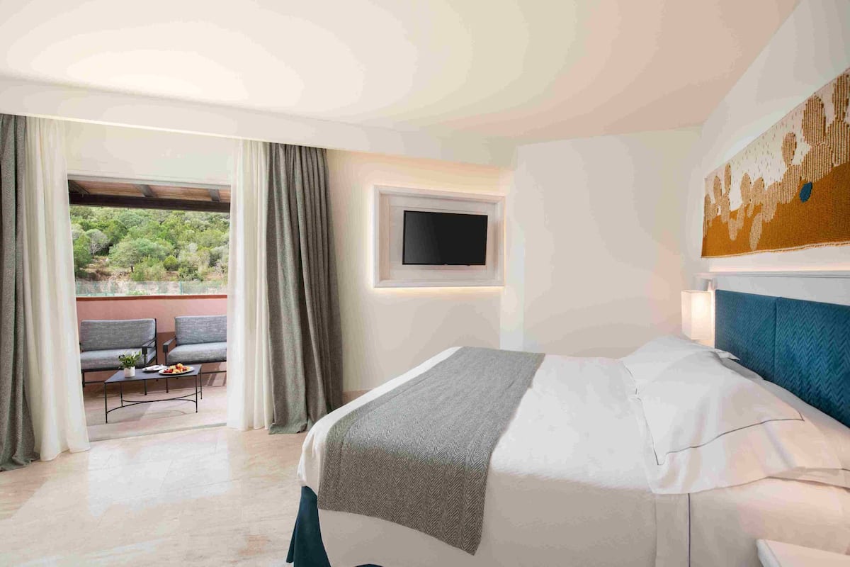 A hotel room with a bed, a TV mounted on the wall, a window with curtains, and a balcony with seating.
