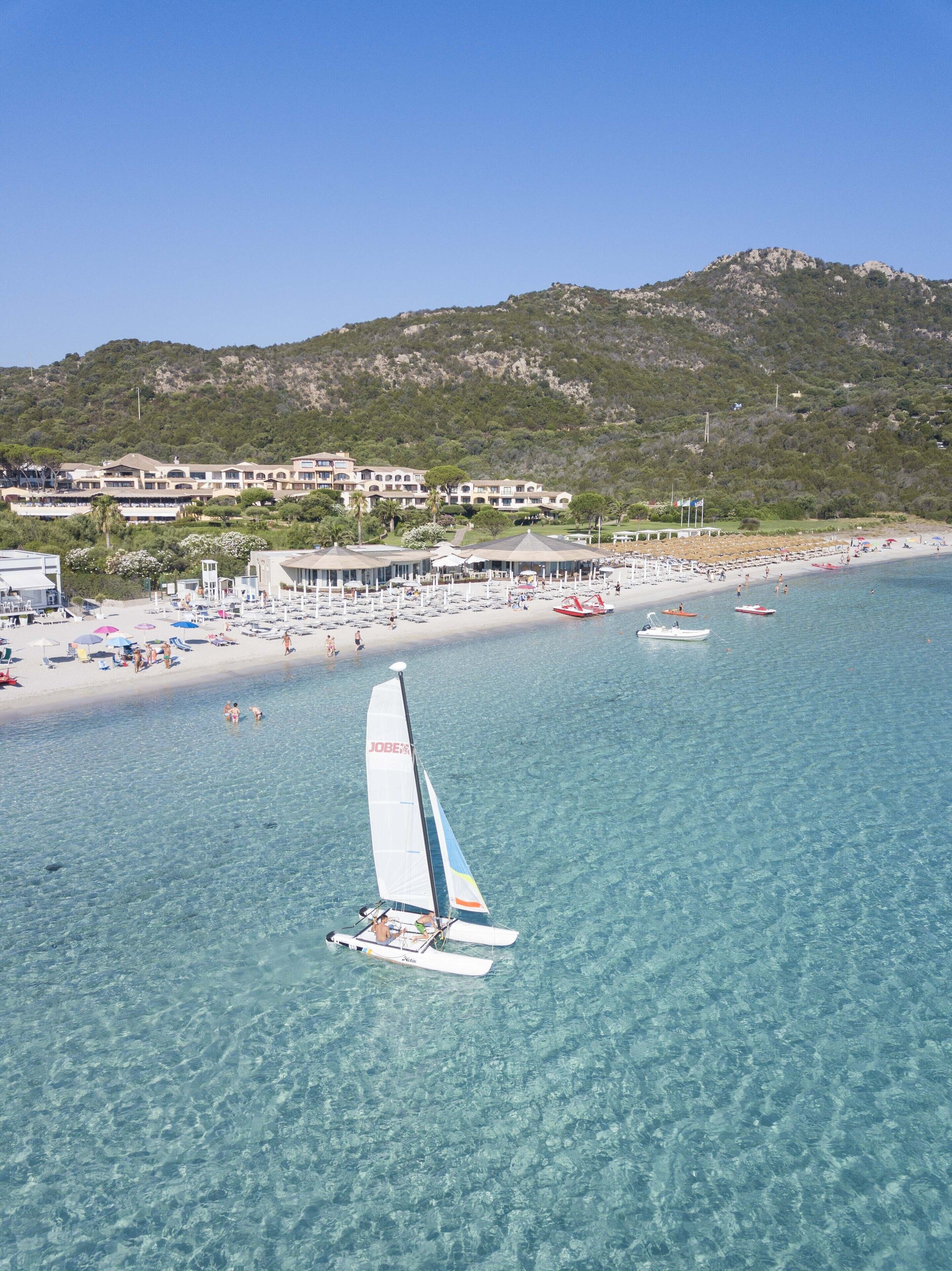 A coastal area with a clear beach, a sailboat, and a building complex in the background.