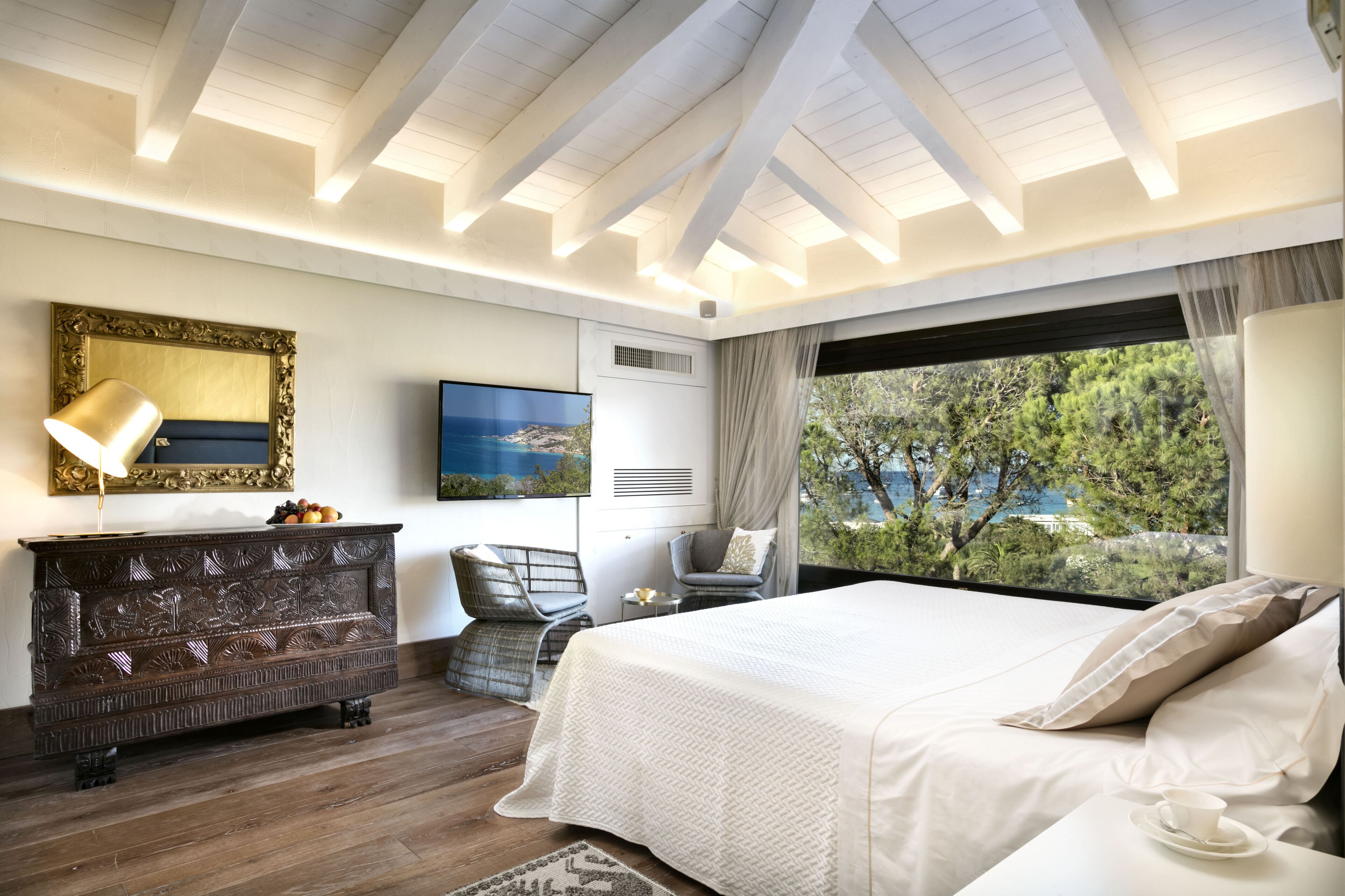 A bedroom with a large bed, a wooden dresser, a chair, and a view of greenery through a window.