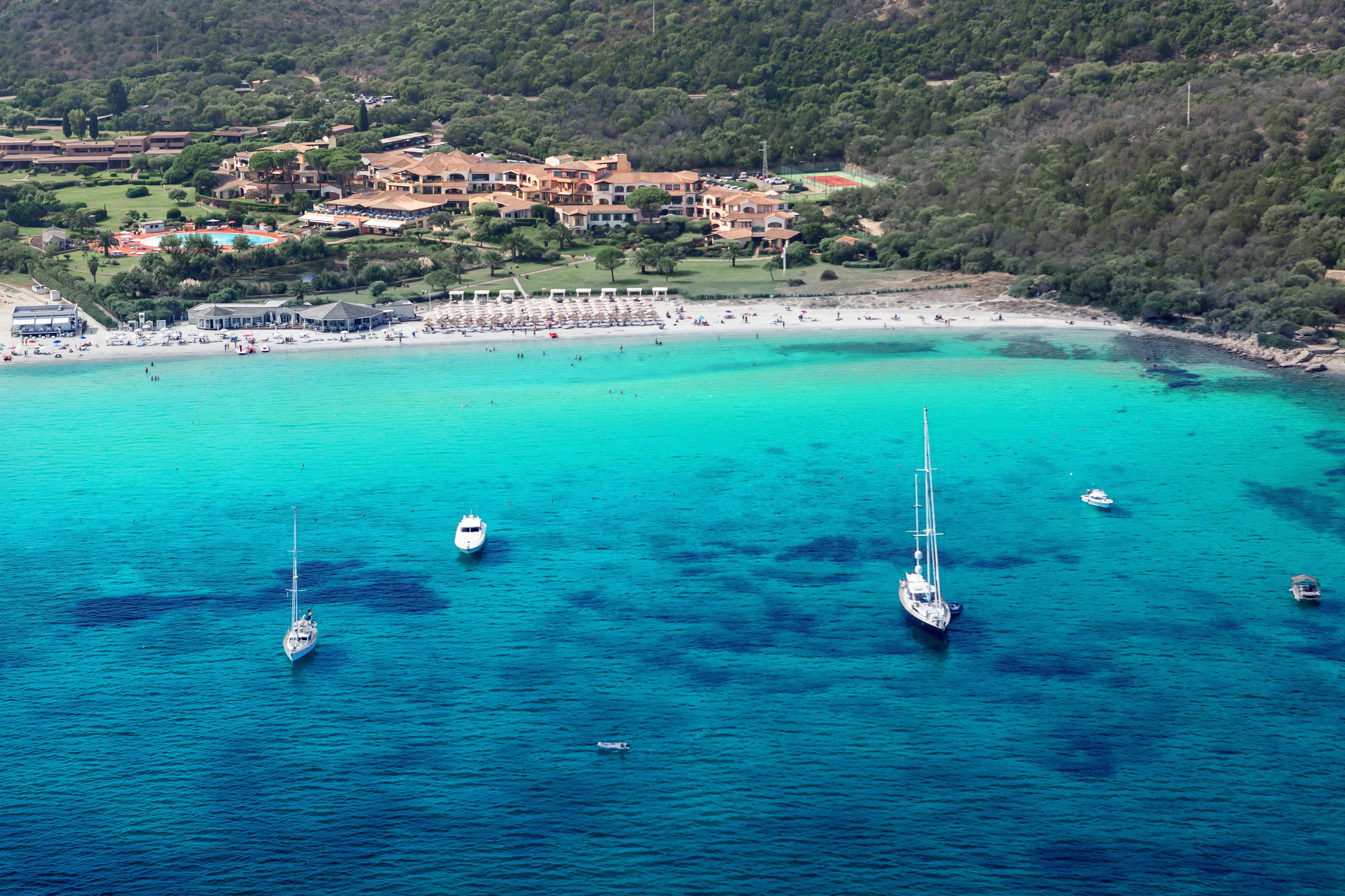 Vue aérienne d’une zone côtière avec une plage, des bateaux et un complexe hôtelier comprenant plusieurs bâtiments.