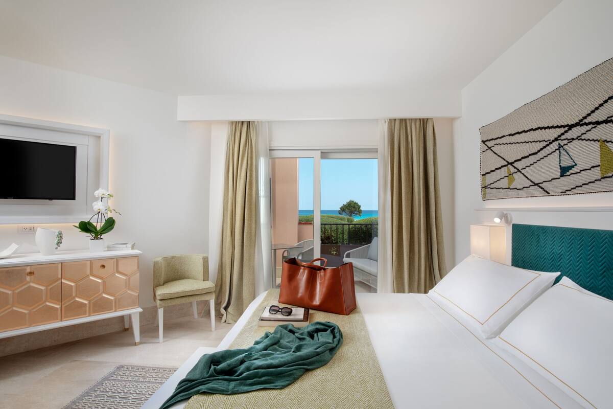 A hotel room with a bed, a TV, a desk, a chair, a window with curtains, and a view of the outdoors.