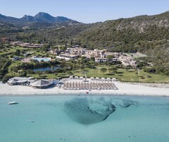 Vista aérea de un complejo turístico costero con varios edificios, una playa con sombrillas y una piscina.