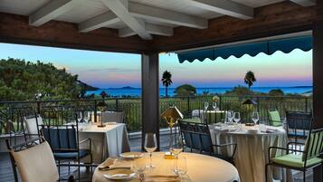Un espace de restauration en plein air avec des tables pour deux, offrant une vue panoramique sur l’océan et les collines.