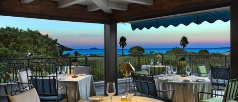 An outdoor dining area with tables set for two, overlooking a scenic view of the ocean and hills.