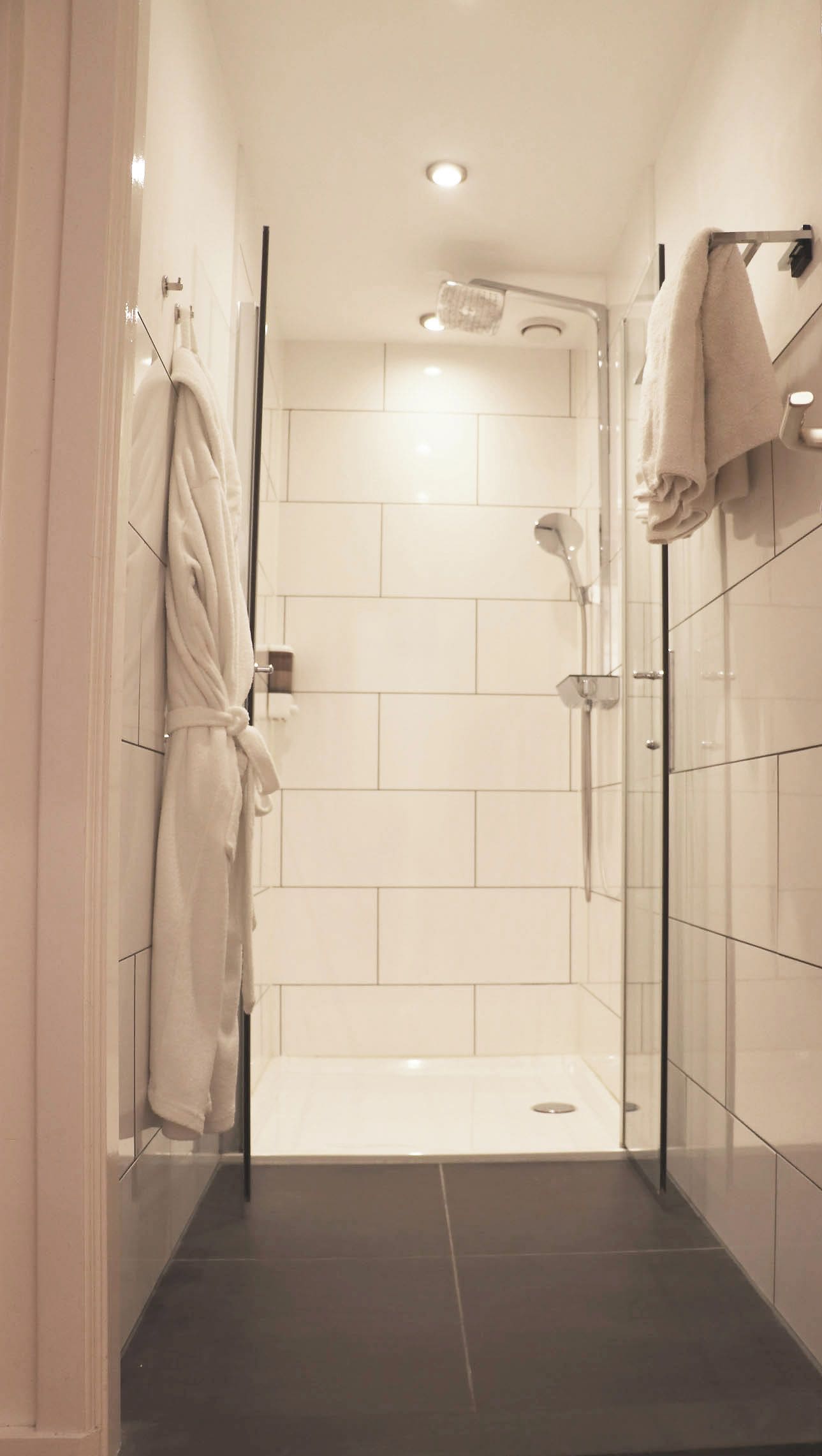 A modern bathroom with a glass shower enclosure, white tiles, and a dark floor.