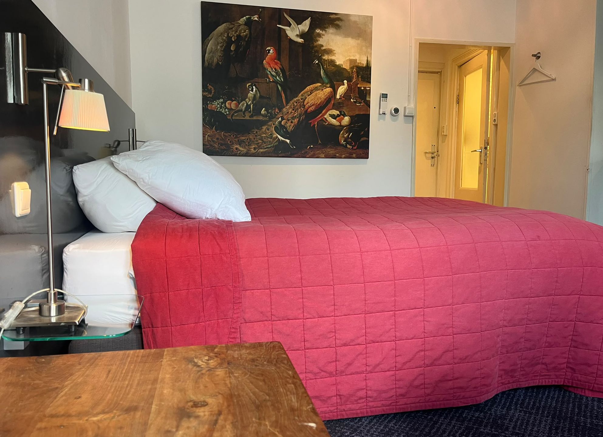 Una habitación de hotel con una cama, una mesita de noche, una lámpara, un cuadro en la pared y una puerta.