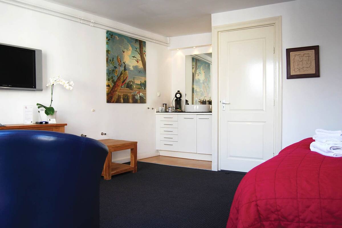 A hotel room with a bed, a desk, a chair, a painting, a mirror, and a door.