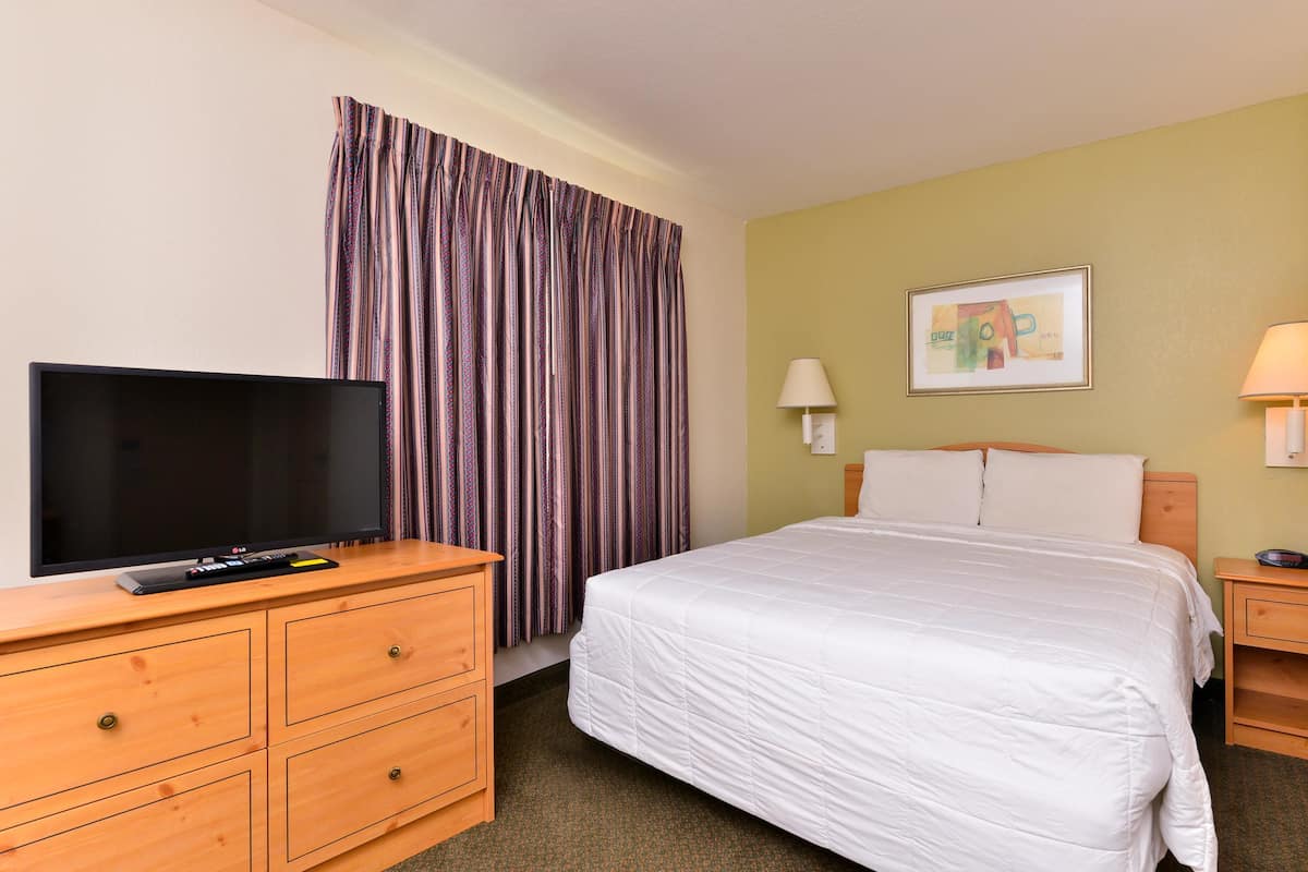 A hotel room with a bed, a TV on a wooden dresser, a lamp, and a painting on the wall.