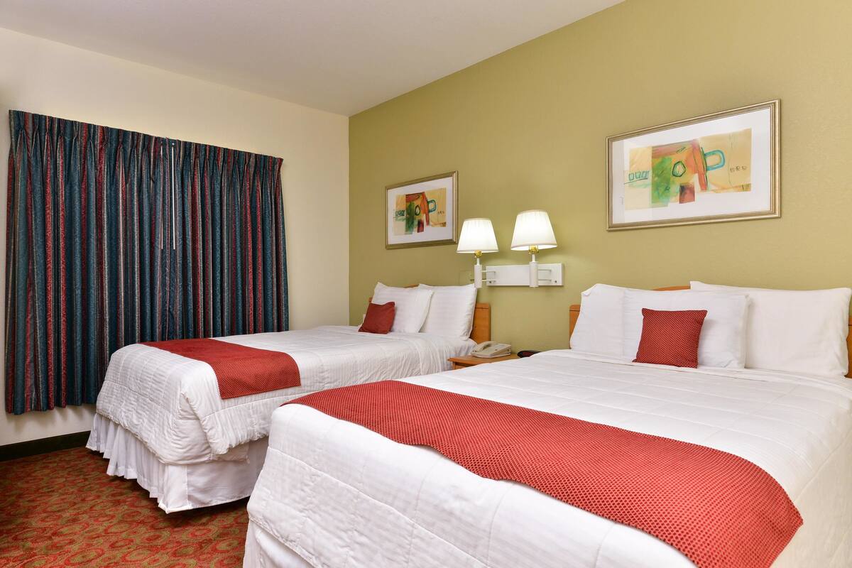 A hotel room with two beds, a red and white checkered carpet, a large window with blue curtains, and two wall-mounted lamps.